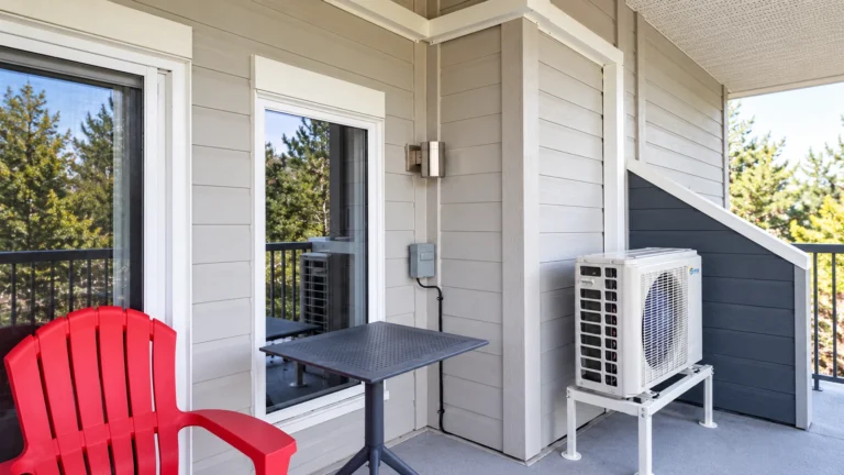 Outdoor mini-split heat pump installed on a raised stand beside a covered residential balcony with patio furniture.